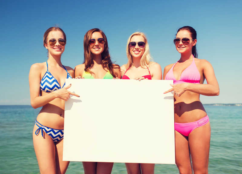 四名比基尼美女在海滩上展示白色的空白广告牌
