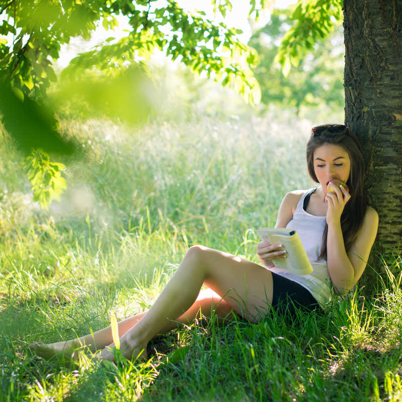 美丽的年轻女子在公园的树下看书