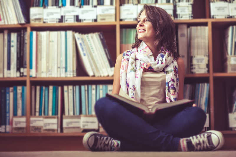 美丽的女孩坐在书架边腿上放着一本书
