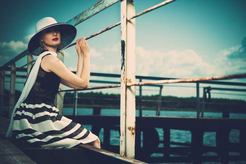美丽的女人戴着帽子和围巾坐在码头上