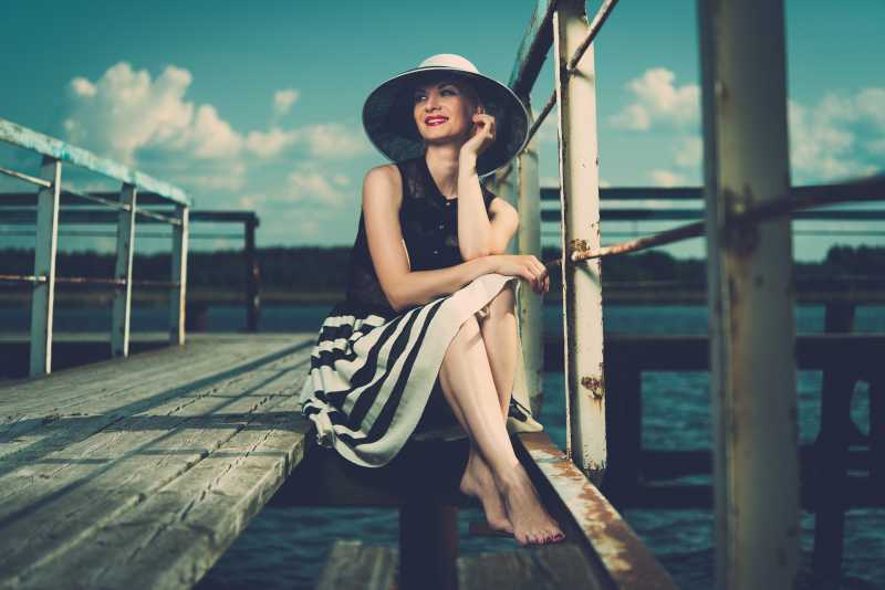 美丽的女人戴着帽子坐在码头边上