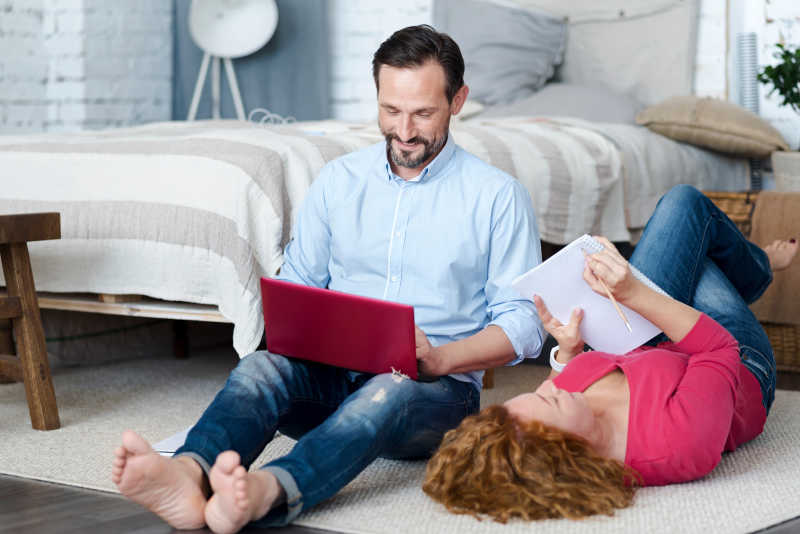 躺在地毯上做笔记的妻子和坐在地毯上使用笔记本电脑的丈夫