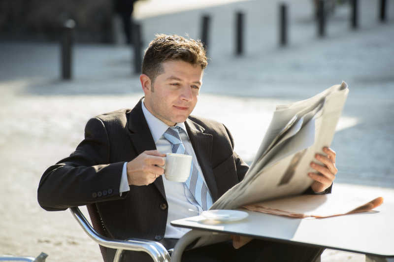 有魅力的商人坐在户外喝咖啡看报纸