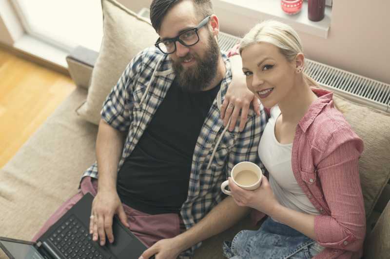 亲密的夫妇坐在沙发上一边使用笔记本电脑一边喝咖啡