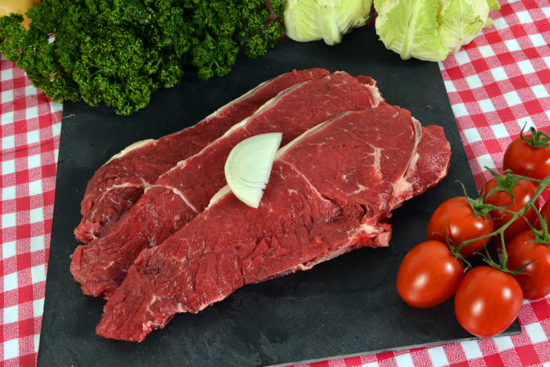 红色格子桌布背景下黑色砧板上的的生牛排和一些蔬菜