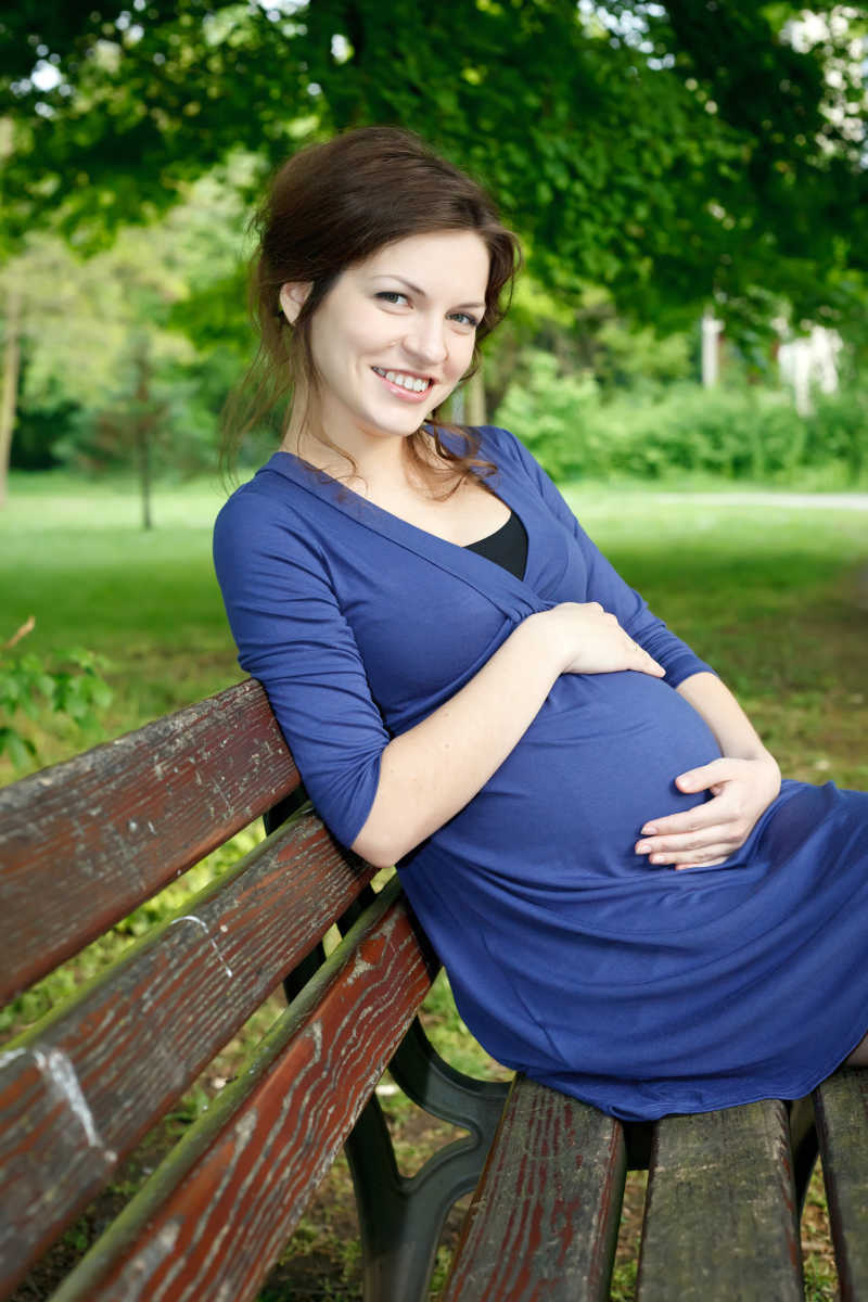 绿色户外背景下坐在长椅上的年轻孕妇