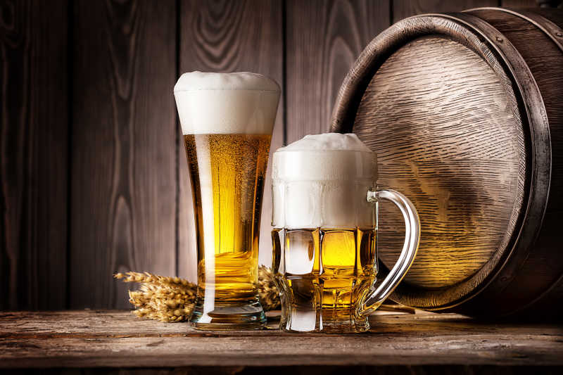 木制圆桶旁边的两杯黄色的泡沫啤酒