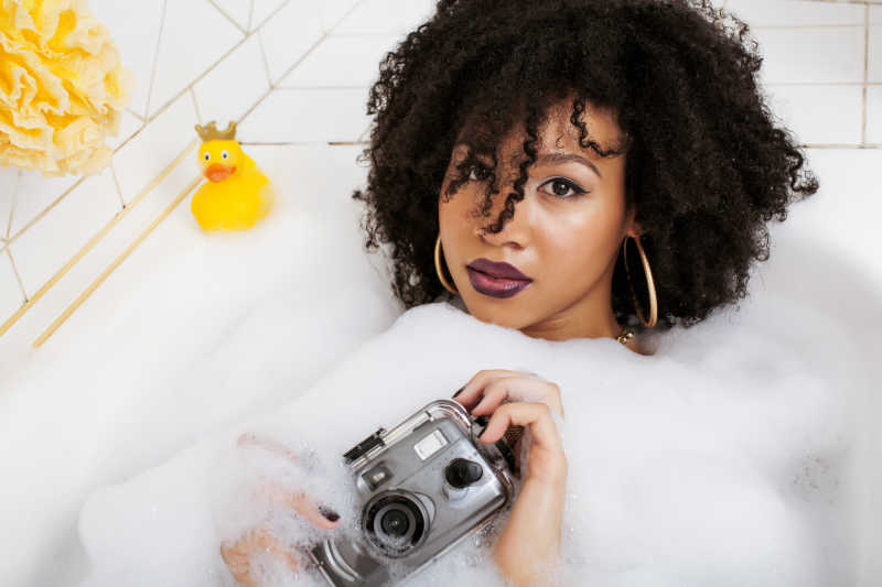 年轻的黑人卷发少女泡在浴缸里拿着相机