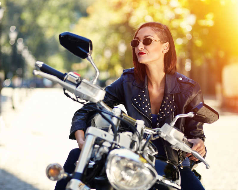 有阳光的绿色植物背景下坐在摩托车上穿着皮夹克的女孩
