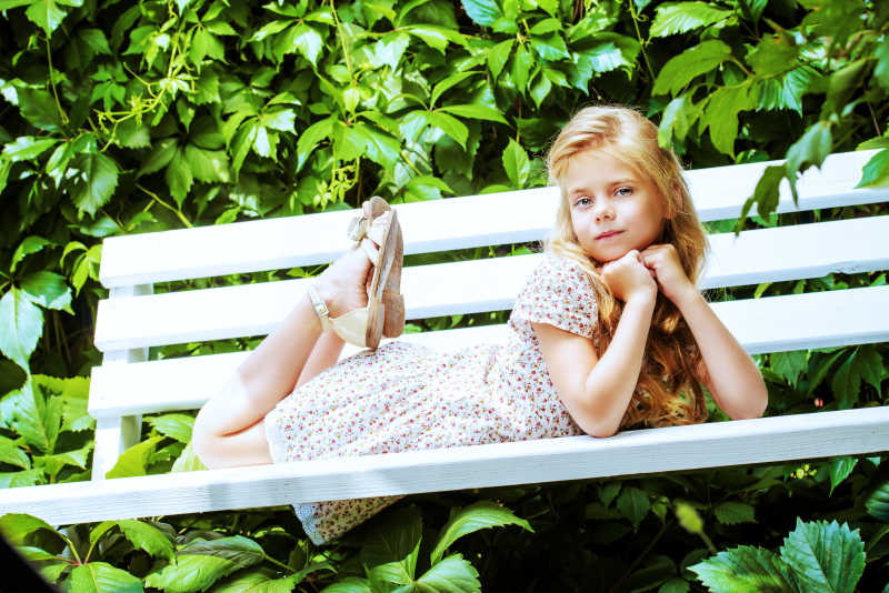阳光下绿色树叶背景下的趴在长椅上的金发碧眼的小女孩