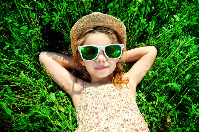 阳光下躺在草地上戴着太阳镜用胳膊枕着头的小女孩