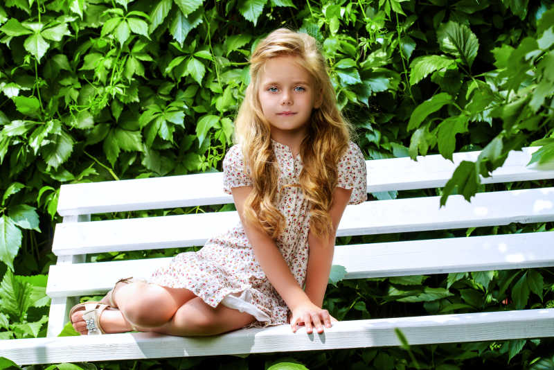 绿色植物背景下坐在白色长椅子上的金发碧眼的小女孩