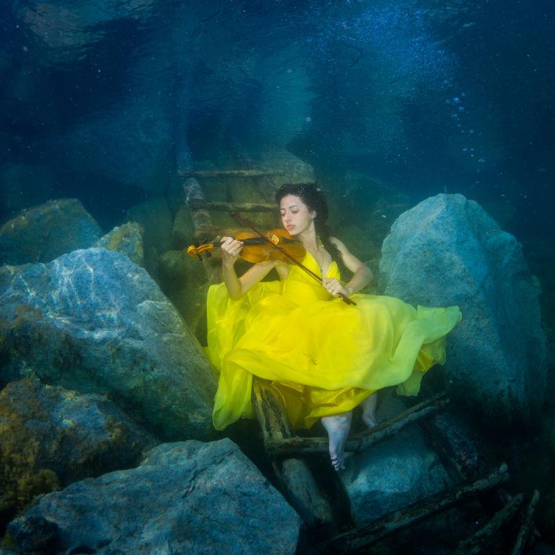 穿着黄色裙子在水下楼梯上拉小提琴的年轻优雅的女子