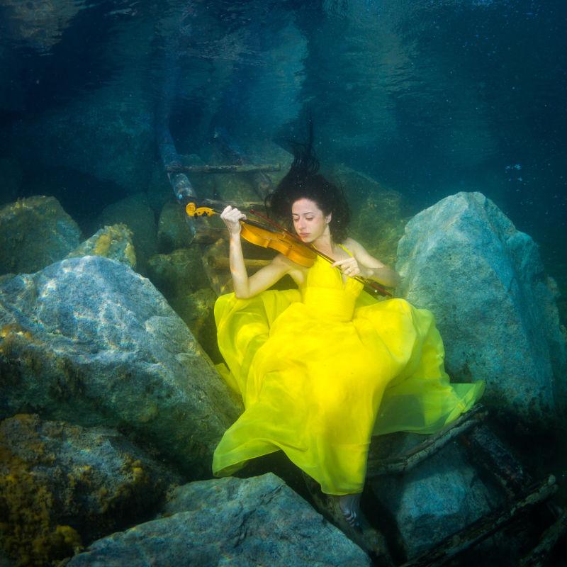 在水下楼梯上穿着一件黄色裙子的年轻的优雅女人拉小提琴