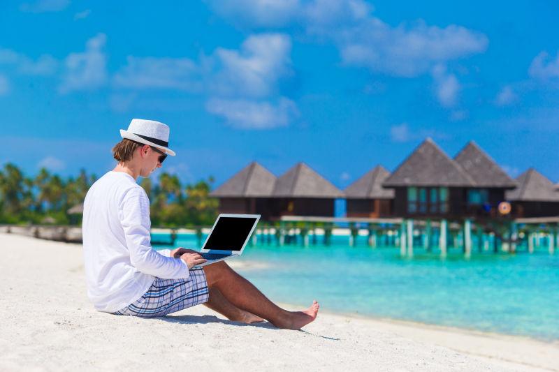 坐在热带海滩上使用笔记本电脑的年轻男人