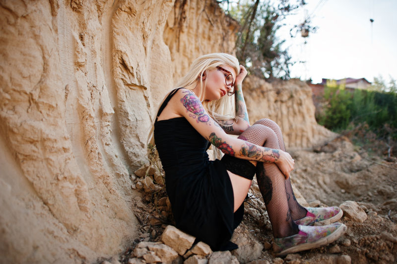 一位纹身金发女郎穿着黑色礼服和长筒袜坐在岩石上