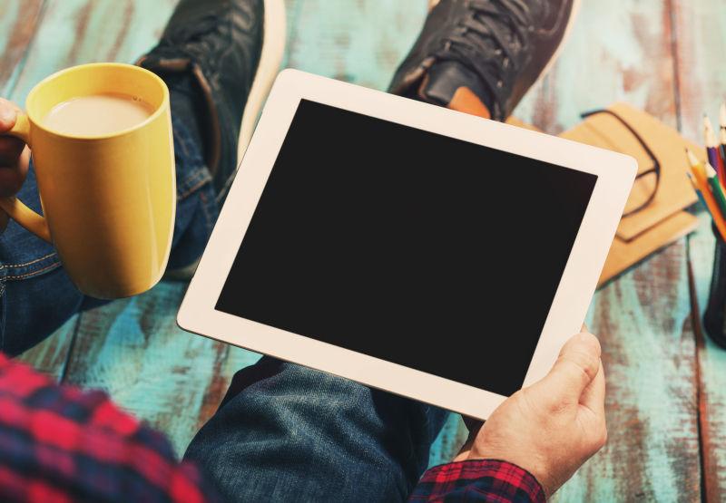 坐在蓝色木地板上一边喝咖啡一边俯视手持数字空间的数码平板