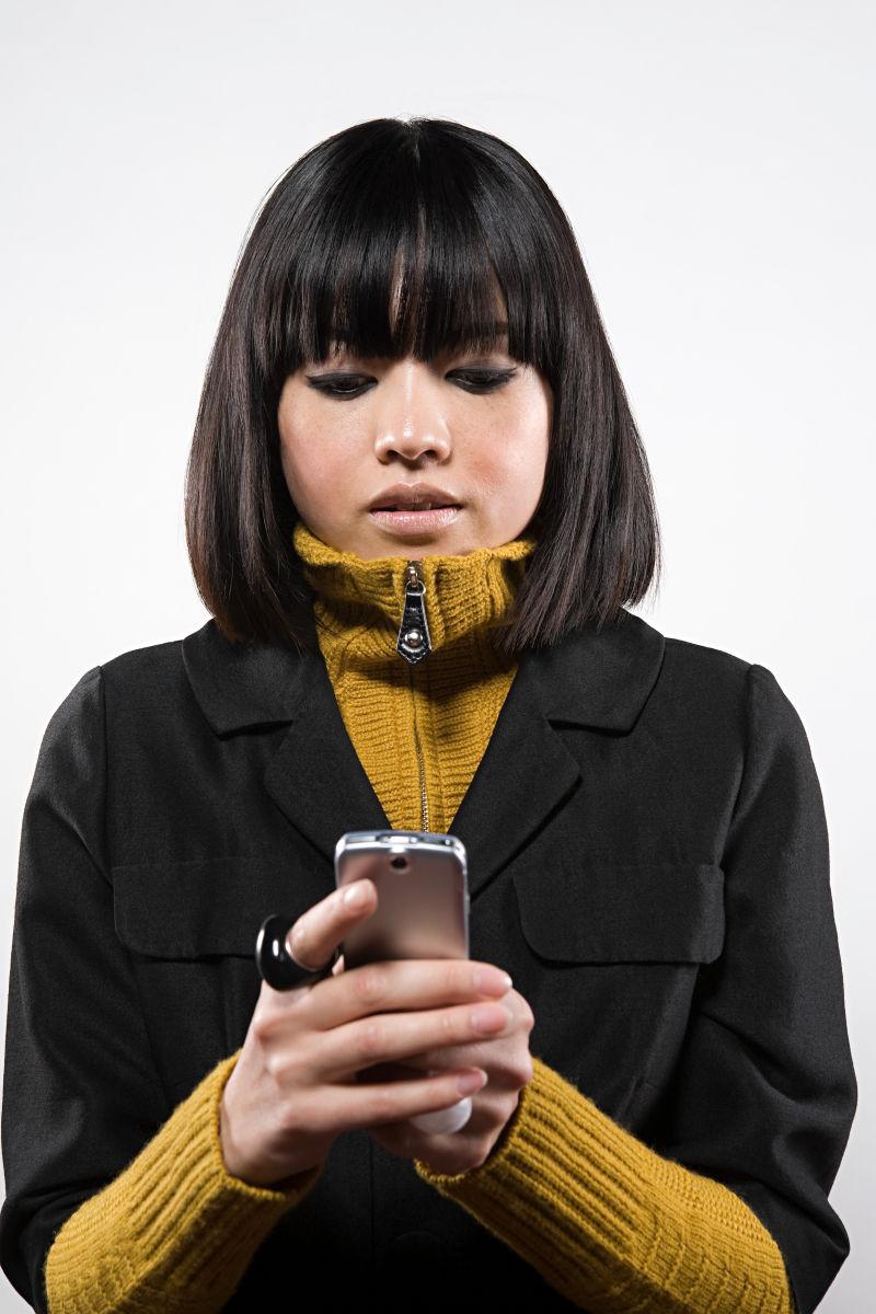 白色背景下穿着黄黑色外套玩手机的亚裔短发美女