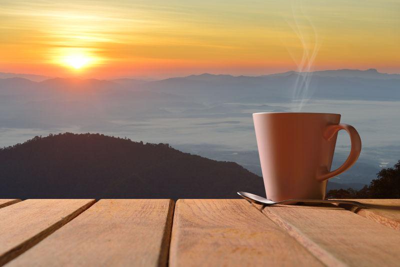 远处山顶上的日出背景下的木桌上的一杯热咖啡和勺子