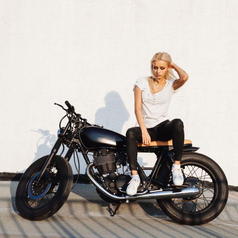 白色背景上阳光下坐在黑色摩托车上的白衣金发美女