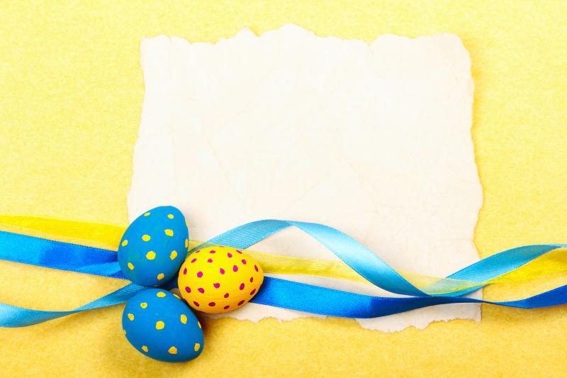 黄色背景上的空白纸片旁的复活节彩蛋和蓝色丝带