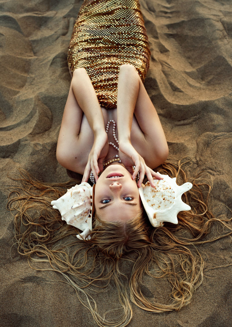 夏末的沙滩上有着金色鳞片巨大的粉红色贝壳和飘逸的头发的美人鱼躺在沙滩上