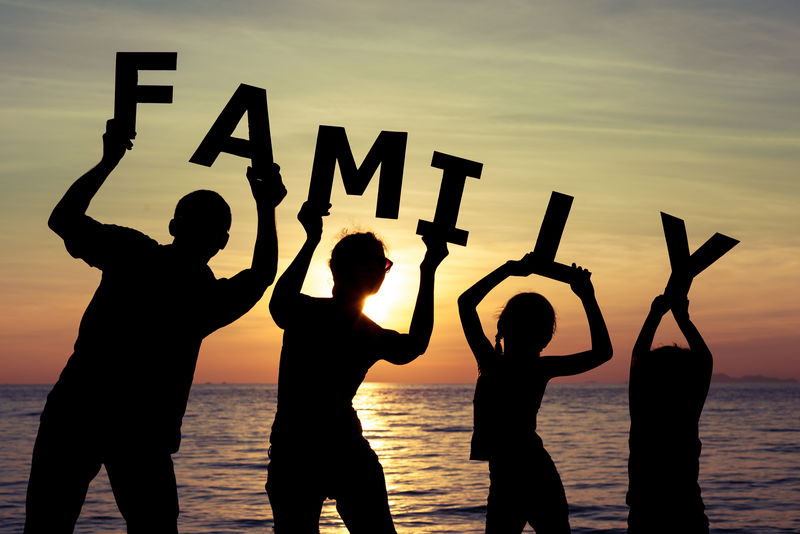 日落时分幸福的一家人站在海滩上-他们保留构成单词“家庭”的字母-友好家庭的概念