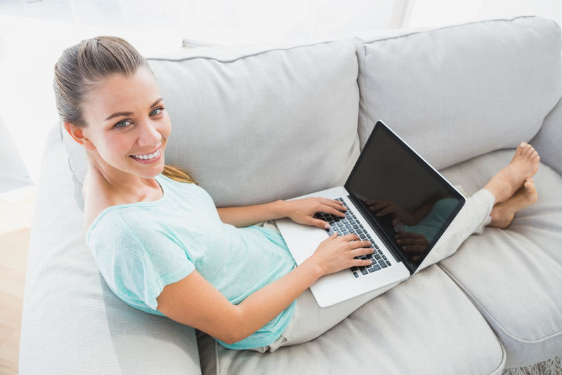 坐在沙发上的快乐女人用笔记本电脑对着客厅里的相机微笑