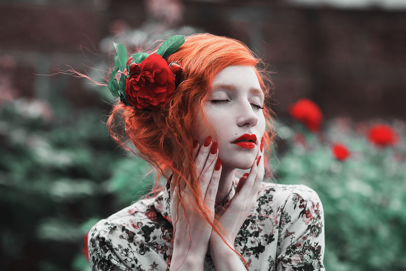 一个穿着花裙的红色卷发女人，背景是一丛红玫瑰。花园里的红发姑娘，皮肤苍白，眼睛蓝，容光焕发，嘴唇红润，腰身纤细。