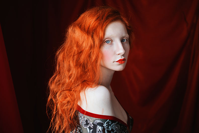 一个女人，红色卷发，穿着红色背景的浴袍。红发姑娘，皮肤苍白，眼睛蓝，容貌异常，嘴唇红润。