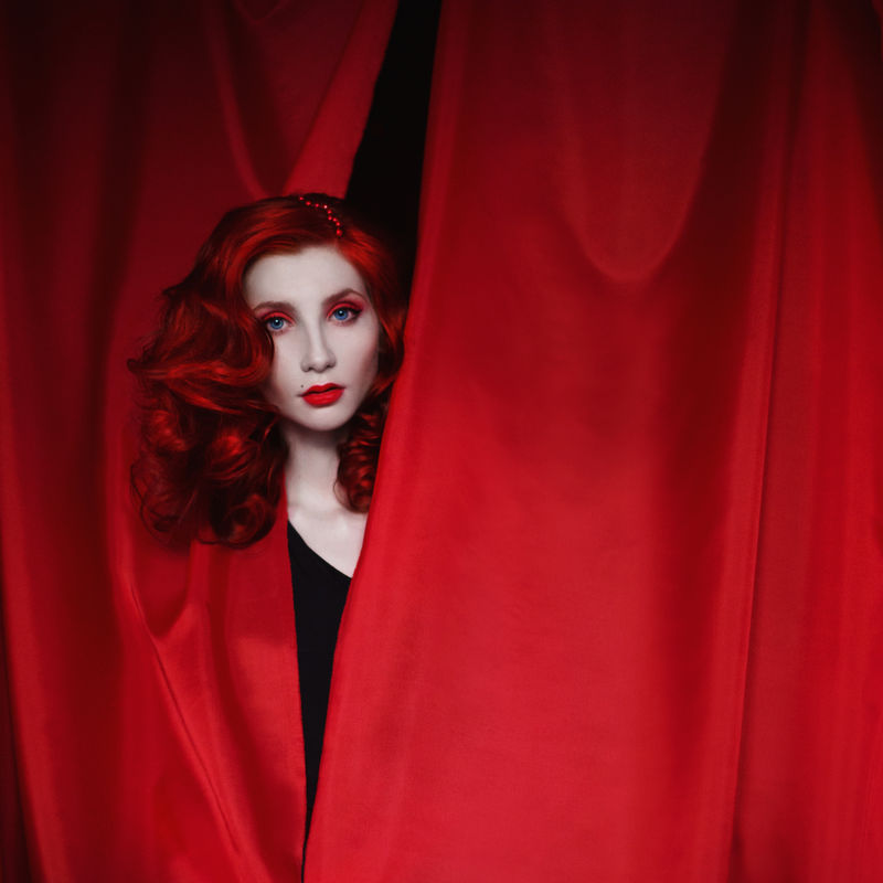 一个穿着黑色连衣裙，留着红色卷发的女人，用红色的布料向外看。红发姑娘，皮肤苍白，腰身细，眼睛蓝，容光焕发，嘴唇红润。复古化妆。戏剧演员