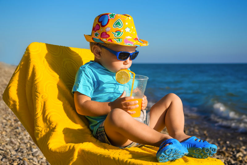 戴着太阳眼镜、戴着帽子的男孩宝宝在沙滩上喝烧杯里的果汁