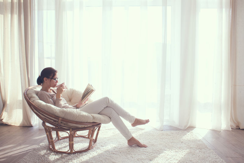 家里的年轻女子坐在窗前的现代椅子上-在起居室里放松地看书-喝咖啡或茶