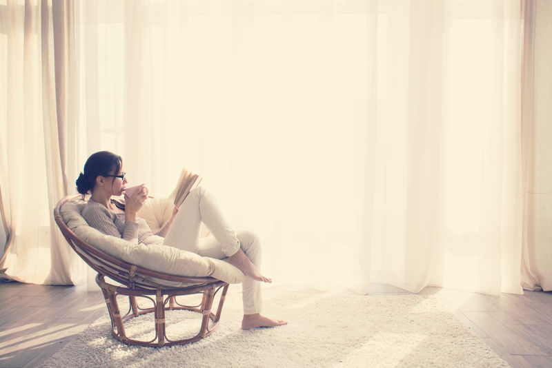 坐在窗户前的现代椅子上的年轻女性在起居室里放松地看书-喝咖啡或茶-喝Instagram Toning