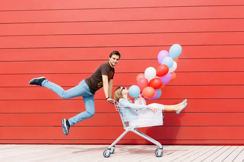快乐的年轻夫妇的照片-女孩坐在购物车里-手里拿着彩色气球-人在跳-红色背景-户外