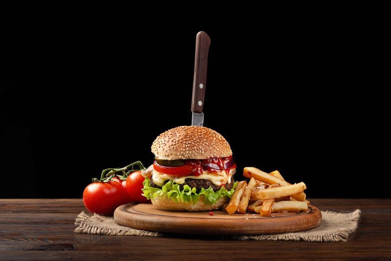 自制汉堡特写镜头与牛肉-西红柿-生菜-奶酪和炸薯条在砧板上-在汉堡里插了一把刀-深色背景下的快餐