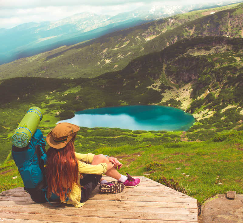 大山和湖泊背景下坐在木桥上休息的女徒步旅行者