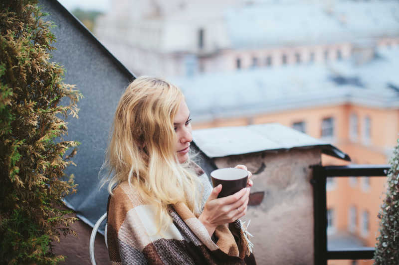 城市背景下坐在阳台上喝咖啡的金发美女